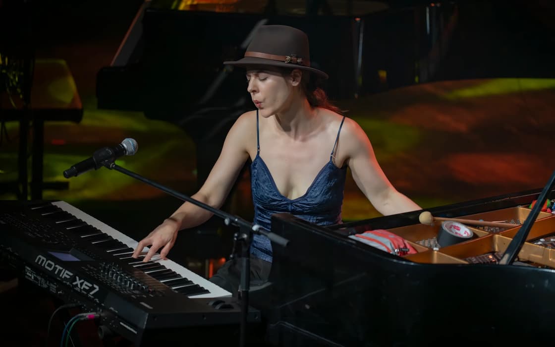 A woman in a hat plays a keyboard and piano on a lit stage.