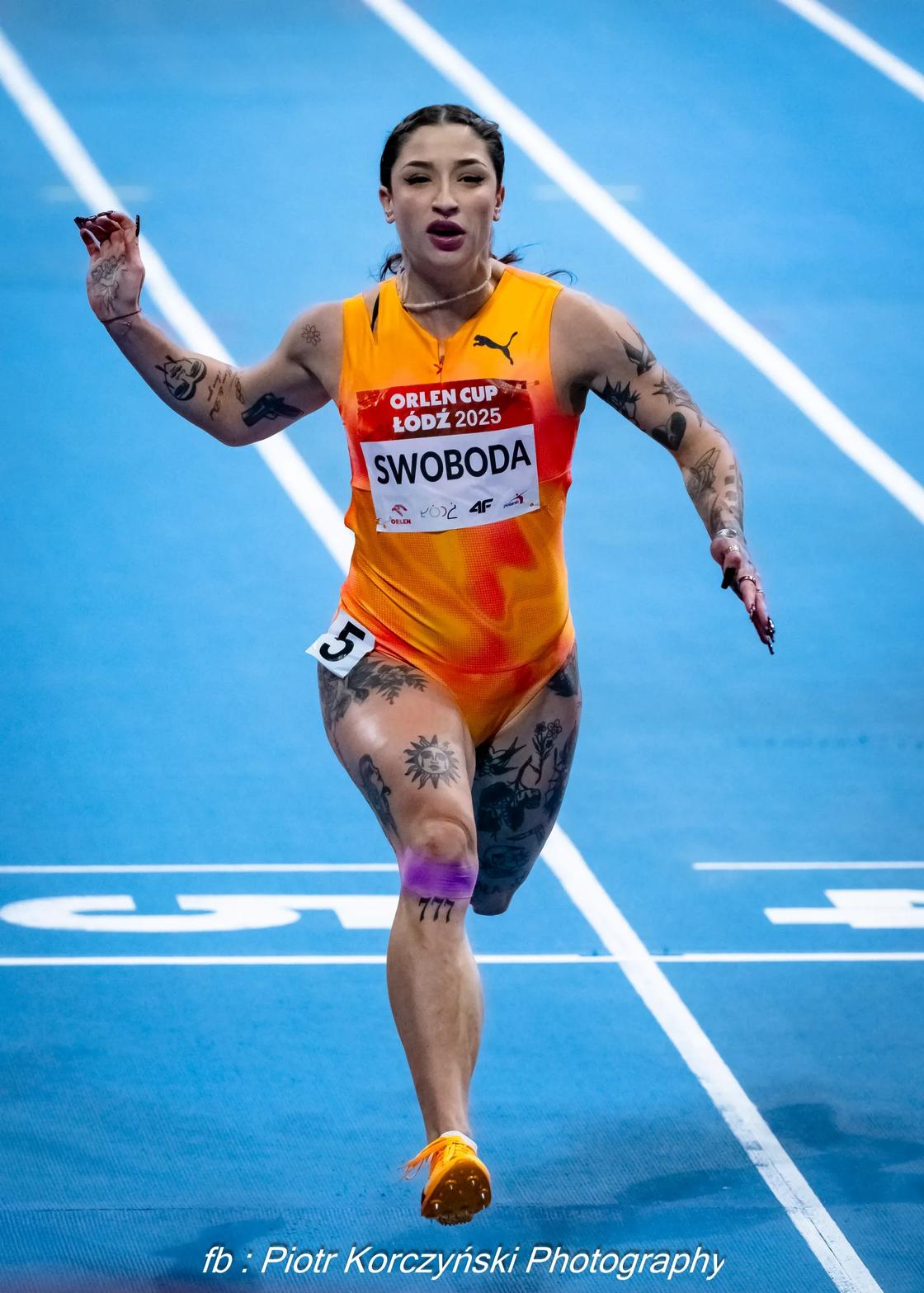 A sprinter in an orange kit races across the finish line on a blue indoor track.