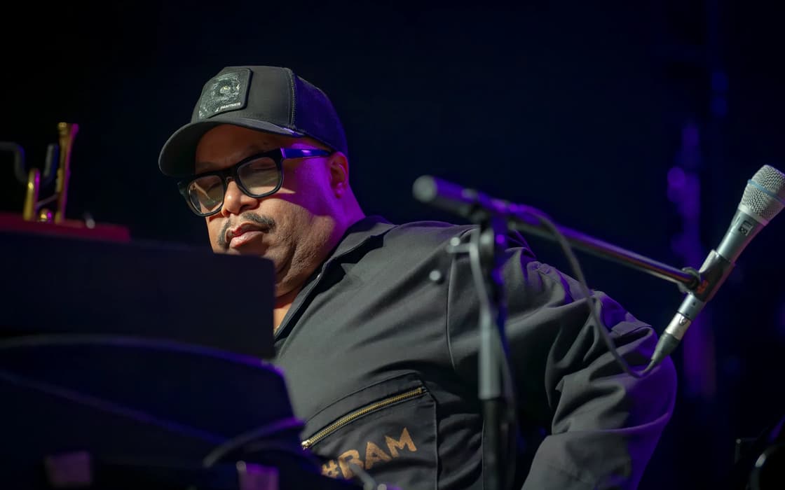 A man in glasses and a cap sits at a piano onstage, with microphones positioned beside him.
