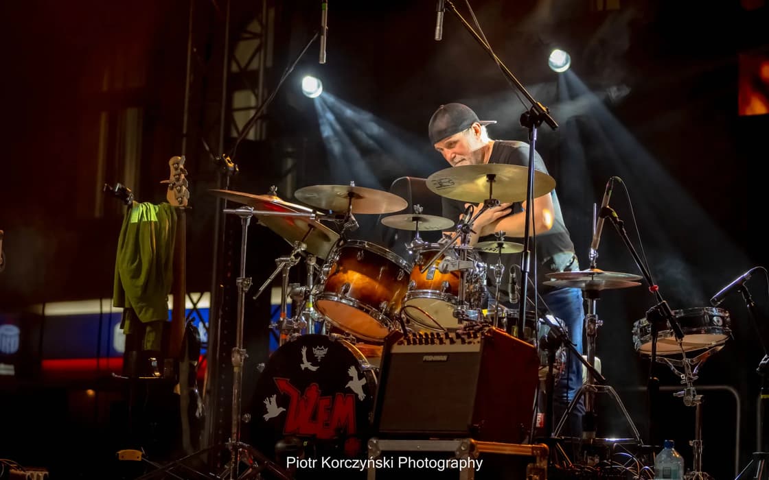 A drummer plays on stage behind a drum kit, lit by spotlights and surrounded by concert equipment.