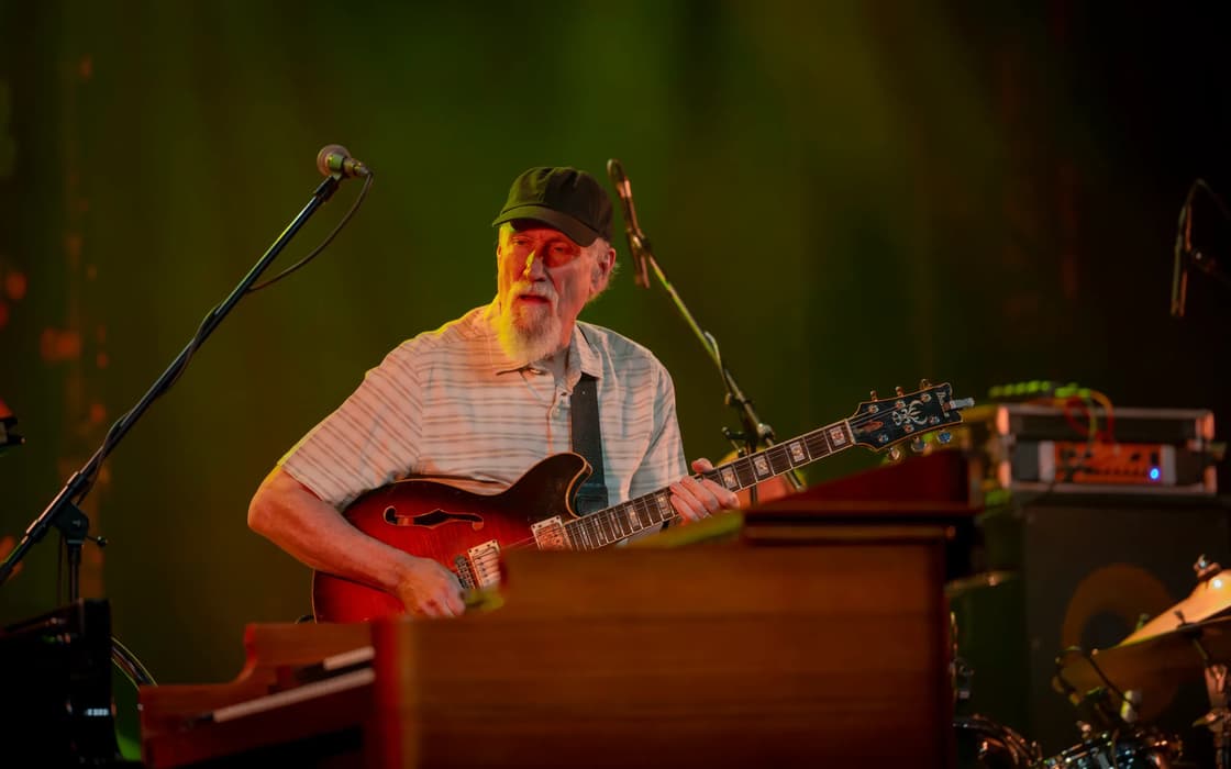 A man in a cap plays electric guitar onstage, partly obscured by a keyboard in the foreground.