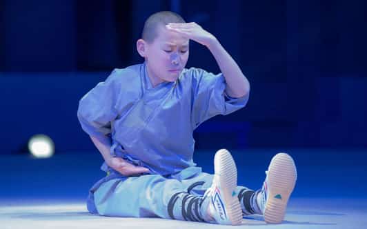 Młody mnich Shaolin siedzi na scenie z wyciągniętymi nogami, dłonią przy brzuchu i drugą przy czole.