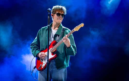 Mężczyzna w okularach gra na czerwonej gitarze przy mikrofonie na scenie z niebieskim oświetleniem.