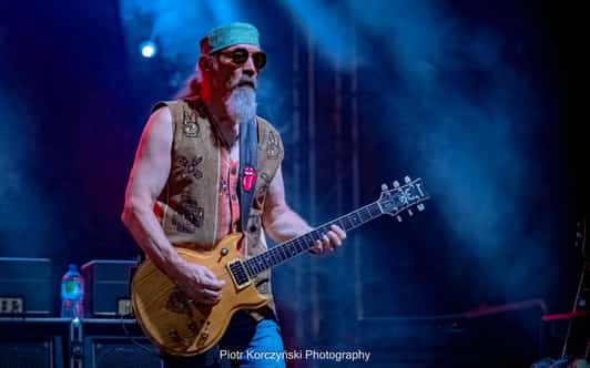 Mężczyzna z długą brodą i gitarą elektryczną stoi na scenie w niebieskim świetle.