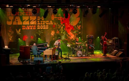 Czterech muzyków gra na scenie jazzowej z fortepianem, gitarą, perkusją i kontrabasem.