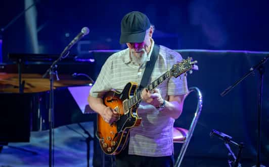 Mężczyzna w czapce gra na gitarze elektrycznej na scenie oświetlonej niebiesko-fioletowym światłem.
