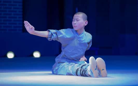 Młody adept Shaolin w szarym stroju wykonuje siedzącą pozycję kung fu na oświetlonej scenie.