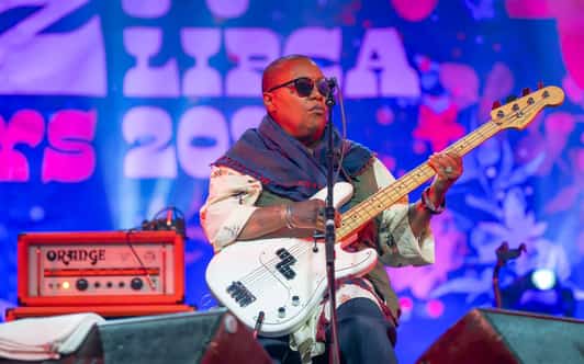 Basista w okularach gra na białej gitarze basowej na kolorowo oświetlonej scenie obok wzmacniacza.