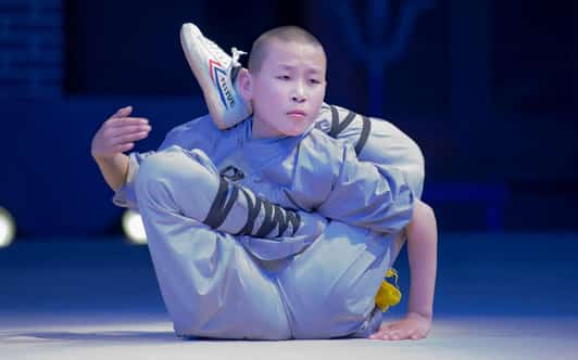 Młody mnich Shaolin w szarej sutannie wykonuje akrobatyczną, kontorsyjną pozę na scenie.
