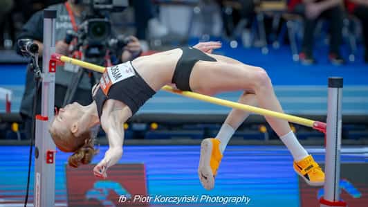 Zawodniczka w czarnym stroju pokonuje poprzeczkę w skoku wzwyż na hali sportowej.