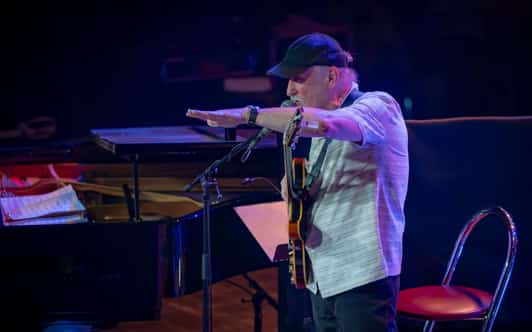 Muzyk z gitarą stoi przy mikrofonie na scenie, unosząc obie dłonie, a obok widać fortepian.