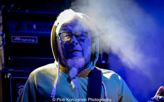 Starszy mężczyzna w bluzie z kapturem i okularach gra na scenie przy niebieskim świetle i dymie.