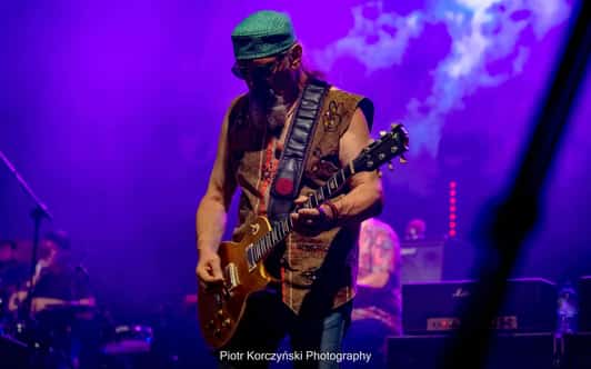 Muzyk w czapce gra na gitarze elektrycznej na scenie skąpanej w fioletowym świetle.