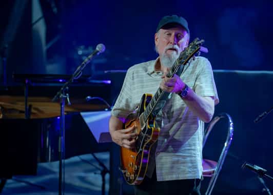 Mężczyzna w czapce gra na gitarze elektrycznej na scenie obok mikrofonu i fortepianu.