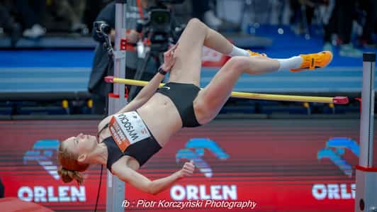 Zawodniczka skacze wzwyż nad poprzeczką na hali lekkoatletycznej.