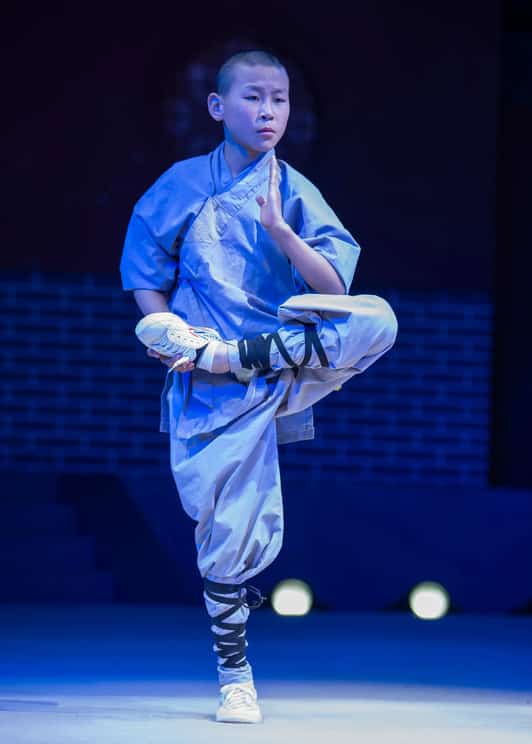 Młody mnich Shaolin stoi na jednej nodze w pozycji kung fu na scenie, ubrany w tradycyjny strój.