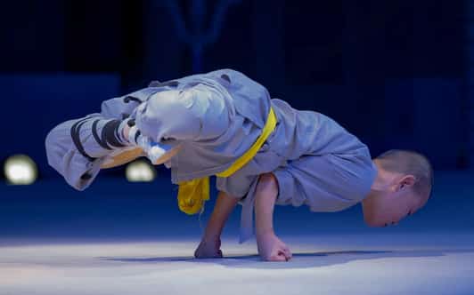 Młody mnich Shaolin w szarej szacie z żółtym pasem balansuje na dłoniach w akrobatycznej pozycji na scenie.