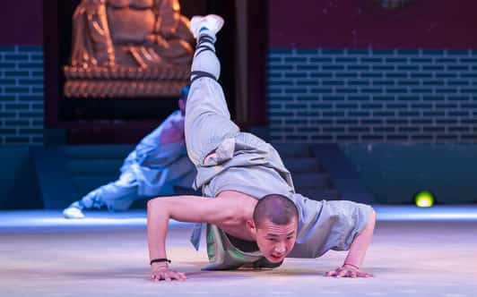 Mnich Shaolin w szarej szacie wykonuje niską podporę jednorącz na scenie przed ołtarzem świątynnym.