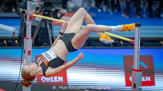 Zawodniczka w czarnym stroju pokonuje poprzeczkę podczas skoku wzwyż na hali lekkoatletycznej.