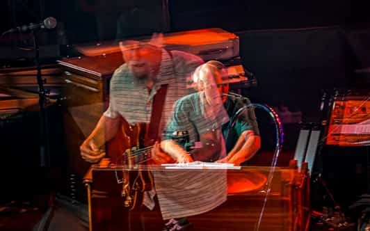Dwaj muzycy na scenie są pokazani z efektem podwójnej ekspozycji, przy gitarze i klawiszach.