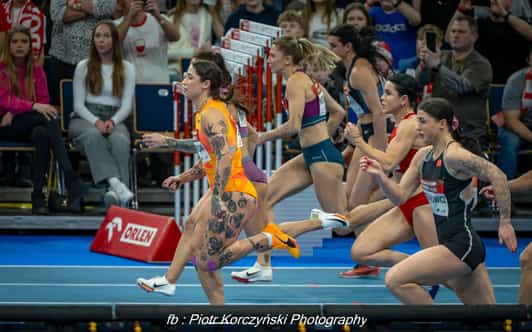 Biegaczki ruszają z bloków startowych na hali lekkoatletycznej, a na trybunach siedzą widzowie.
