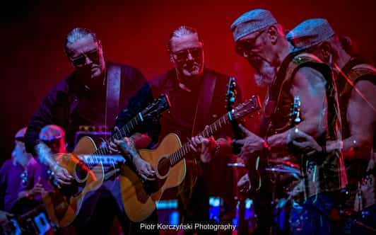 Dwóch gitarzystów gra na scenie w czerwonym świetle, a podwójna ekspozycja pokazuje nakładające się sylwetki.