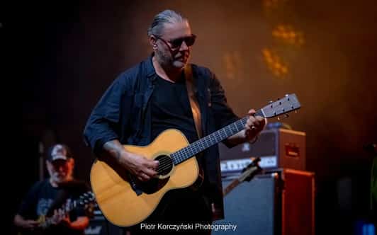 Mężczyzna w okularach przeciwsłonecznych gra na gitarze akustycznej na scenie, a w tle widać drugiego muzyka.