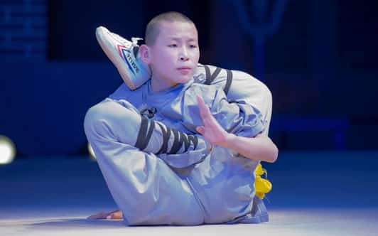 Młody mnich Shaolin w szarej szacie wykonuje skomplikowaną akrobatyczną pozę na scenie.