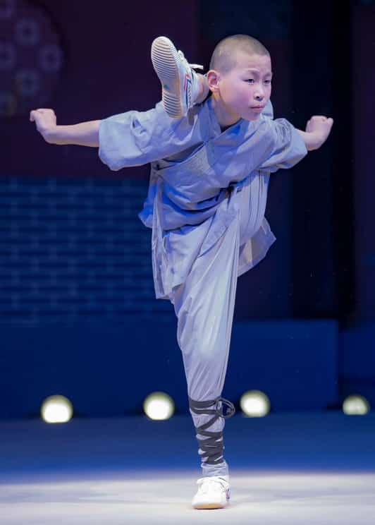 Młody mnich Shaolin w szarej szacie wykonuje wysoki kop balansując na jednej nodze na scenie.