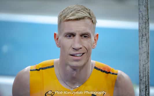 Mężczyzna w żółtej koszulce sportowej stoi przy słupku na rozmytym tle hali lekkoatletycznej.