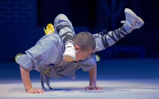 Mnich Shaolin w szarej szacie balansuje na dłoniach na scenie, nogi zgięte nad plecami.