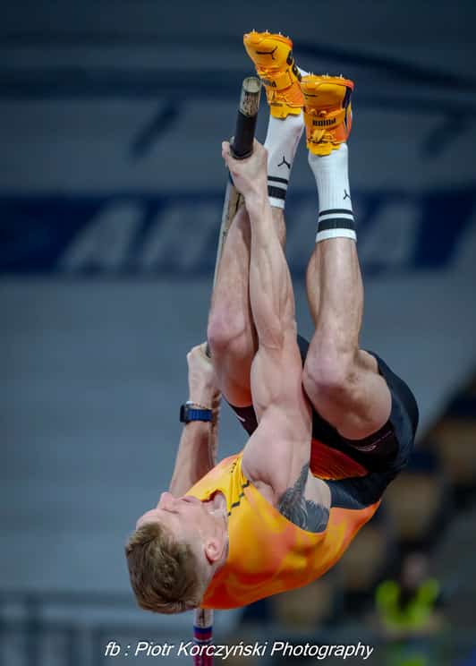 Tyczkarz w pomarańczowym stroju odwraca się głową w dół podczas skoku o tyczce w hali.