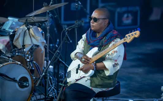 Basista siedzi na scenie z gitarą basową obok zestawu perkusyjnego.