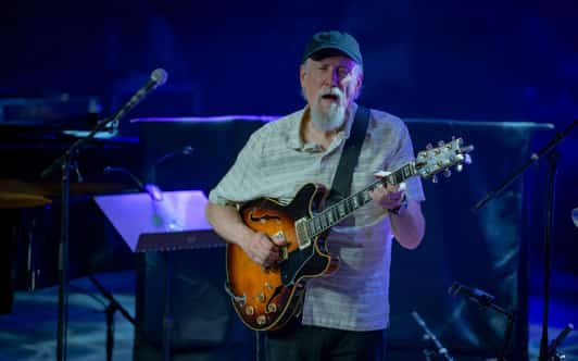 Mężczyzna gra na gitarze elektrycznej na scenie koncertowej w niebieskim świetle.