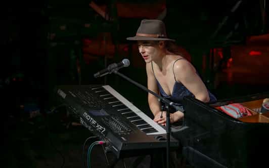 Kobieta w kapeluszu siedzi przy keyboardzie i mówi do mikrofonu na ciemnej scenie.