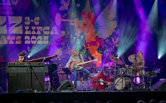 Trzech muzyków gra na scenie z fortepianem, gitarą basową i perkusją na tle festiwalowej scenografii.