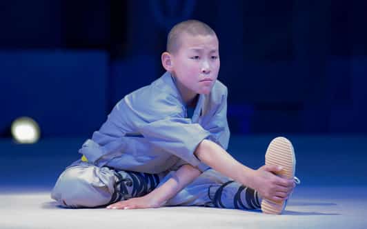 Młody mnich Shaolin w szarym stroju siedzi na oświetlonej scenie i rozciąga nogę, chwytając stopę.