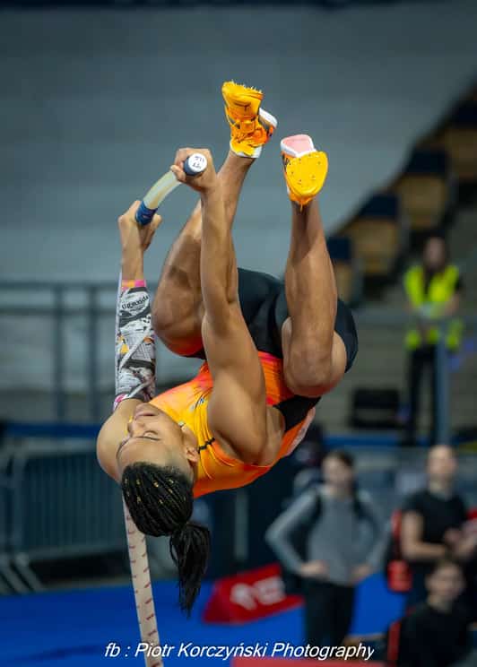 Zawodniczka skacze o tyczce nad poprzeczką w hali sportowej, odwrócona głową w dół.