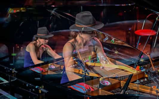 Pianistka w kapeluszu gra przy otwartym fortepianie na scenie, uchwycona z odbiciem w błyszczącej obudowie.