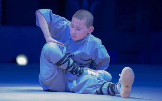 Młody mnich w szarym habicie wykonuje siedzącą pozycję kung fu na niebiesko oświetlonej scenie.