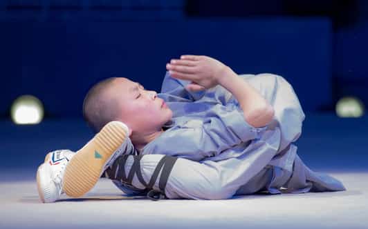 Młody mnich Shaolin w szarej szacie leży na plecach ze stopą przy głowie i złożonymi dłońmi.