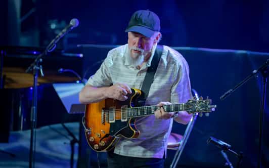 Mężczyzna w czapce gra na gitarze elektrycznej na scenie oświetlonej niebieskim światłem.