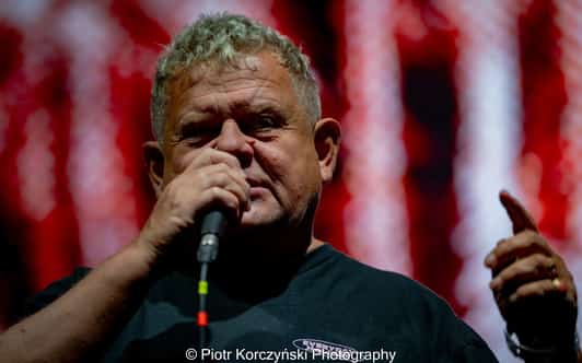 Starszy mężczyzna trzyma mikrofon przy ustach i gestykuluje podczas występu na tle czerwonych świateł.