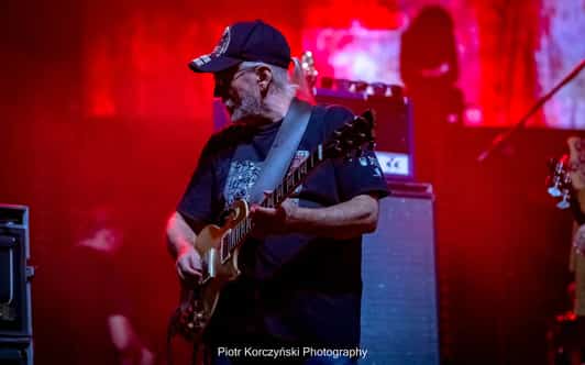 Mężczyzna gra na gitarze elektrycznej na scenie w czerwonym świetle koncertowym.