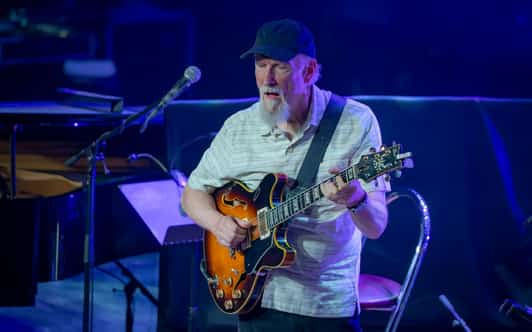 Mężczyzna w czapce gra na gitarze elektrycznej na scenie oświetlonej niebieskim światłem.