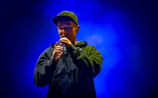 Raper w czapce trzyma mikrofon oburącz na scenie oświetlonej niebieskim światłem.