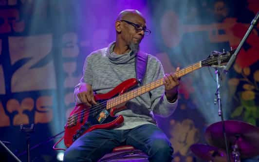 Muzyk siedzi na stołku i gra na czerwonej gitarze basowej na scenie oświetlonej fioletowym światłem.