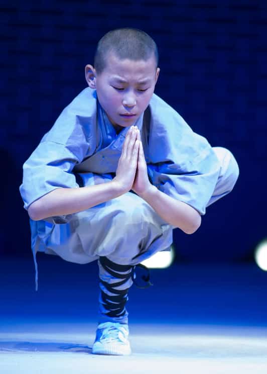 Młody uczeń Shaolin w szarej szacie balansuje w jednonogim przysiadzie z dłońmi złożonymi do modlitwy na scenie.
