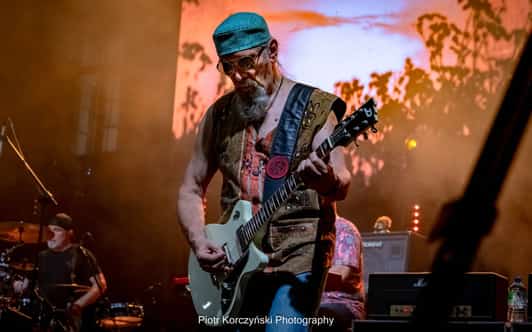 Brodaty gitarzysta w okularach gra na scenie w ciepłym świetle, a w tle widać perkusistę.