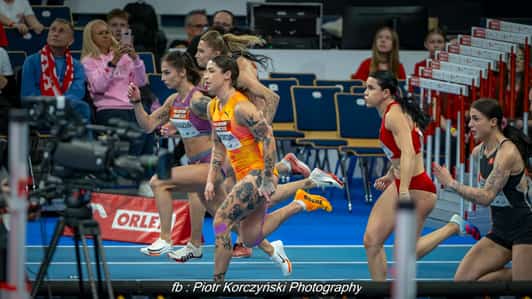 Sześć zawodniczek rusza z bloków startowych na bieżni hali lekkoatletycznej, a z przodu widać kamerę.